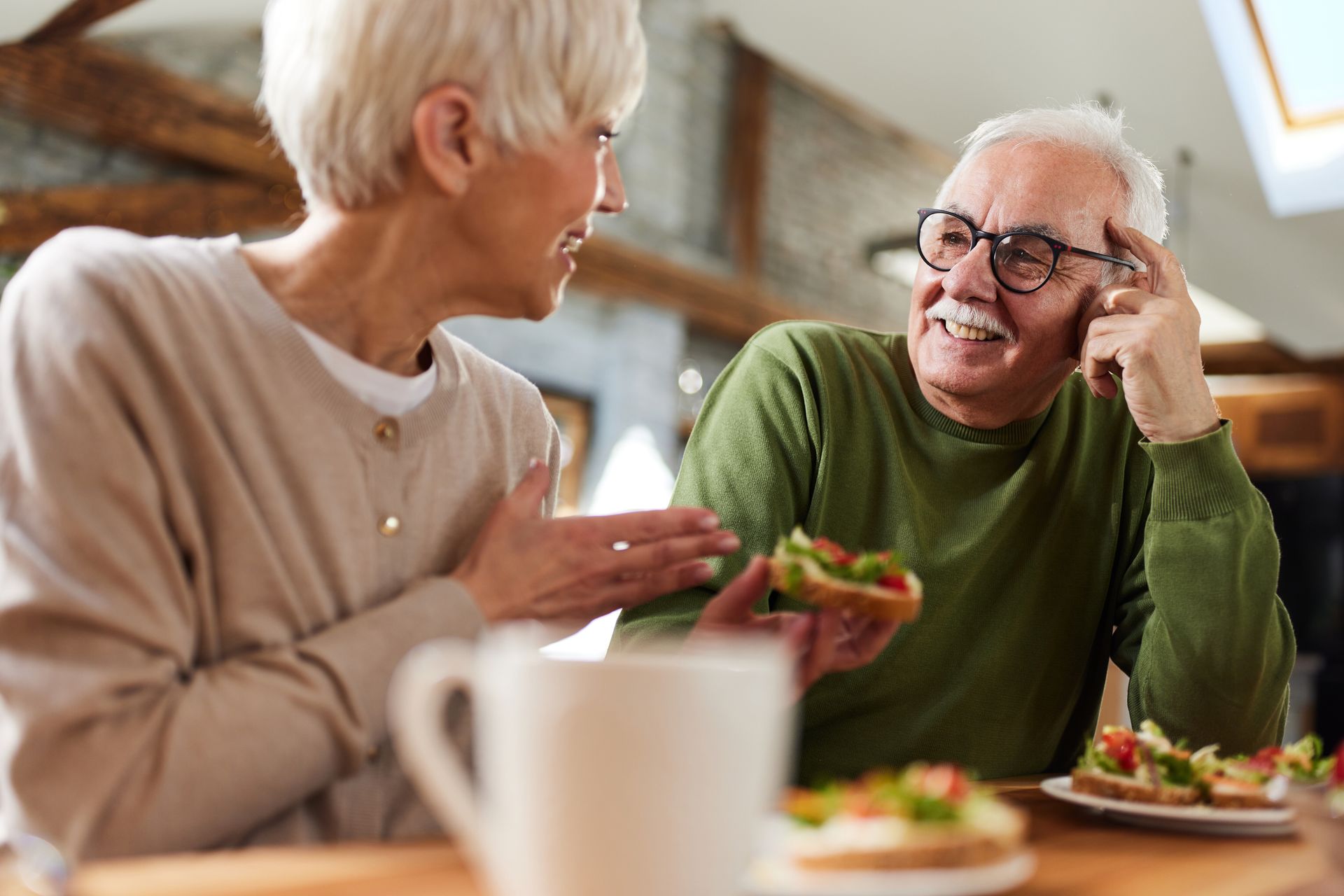 An older couple chatting warmly over lunch at a sunlit kitchen table